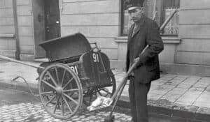 Ein Straßenreiniger um 1906 steht mit Besen und Schaufel vor einer Kehrichtkarre